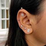 A close-up of a person's ear showcasing a stack of five silver ear piercings, including a dome stud on the lobe, a snowflake, a star, a leaf, and a palm leaf, worn with dark hair.